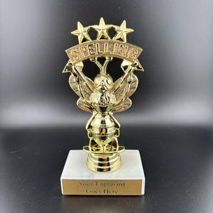May include: A gold-colored spelling bee trophy with a bee figure and the word "SPELLING" above it. The trophy has a white marble base with a gold plate that reads "Your Engraving Goes Here".