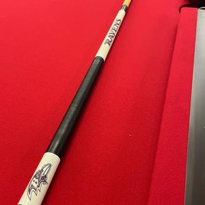 May include: A pool cue with a black grip and white accents. The cue features the word "Ravens" in purple and white, along with a team logo. The cue is set against a red felt surface.