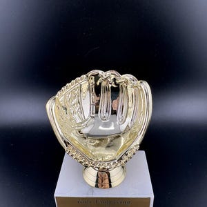 May include: A gold-colored baseball glove trophy on a white marble base. The trophy has a detailed design of a baseball glove and a gold-colored stem. The base has a gold plate with the text "Your Engraving Goes Here".