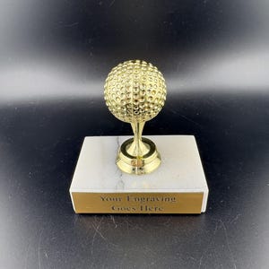 May include: A gold-colored golf ball trophy mounted on a white marble base. The base features a rectangular gold plaque with the black text "Your Engraving Goes Here" centered on the front. The golf ball has a textured dimpled surface.