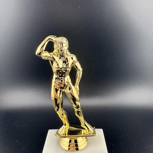 May include: A gold-colored trophy featuring a muscular figure in a bodybuilding pose, standing on a white marble-style base with a metal plate that reads "Your Engraving Goes Here" in black text.