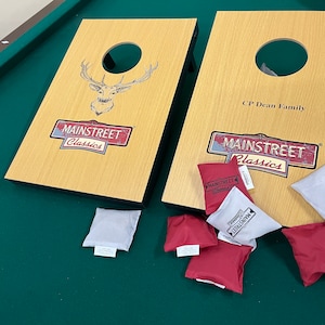 May include: Two wooden cornhole boards with a brown wood grain finish. The boards have a circular hole in the center and are decorated with a deer silhouette and the text "Mainstreet Classics". The boards are on a green felt surface with six bean bags, two white and four red. The red bean bags have the text "Mainstreet Classics" printed on them.