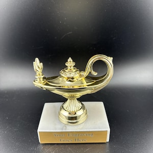 May include: A gold-colored trophy shaped like a genie lamp, atop a white marble base with a gold plaque. The plaque reads "Your Engraving Goes Here". The lamp features a curved handle and a decorative spout.