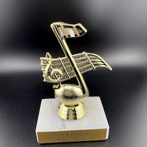 May include: A gold-colored music-themed trophy with a treble clef and musical notes. The trophy sits on a white marble base with a gold plate that reads "Your Engraving Goes Here". The background is black.