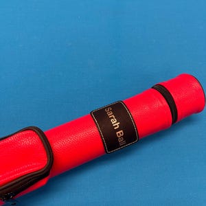 May include: A bright pink pool cue case with a black zipper and a black leather patch that says "Sarah Ball" in silver lettering.