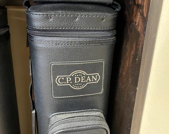 May include: Black leather cue case with a zippered top and a small zippered pocket. The case has a handle and a white rectangular patch with the text "C.P. DEAN" and "ESTD. 1888 COMPANY". The case is designed to hold pool cues.