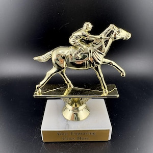May include: A gold-colored trophy featuring a horse and jockey in mid-race. The trophy sits atop a white marble base with a gold-colored pedestal and a rectangular plate that reads "Your Engraving Goes Here."