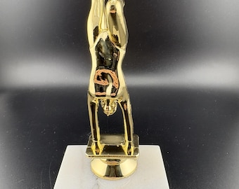 May include: A gold-colored trophy featuring a stylized figure in a handstand pose. The trophy is mounted on a white marble base with a gold-colored plate that reads "Your Engraving Goes Here".