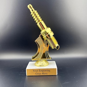 May include: A gold-colored trophy shaped like a spark plug, mounted on a white marble base with a gold plaque. The plaque reads "Your Engraving Goes Here". The trophy is likely for an automotive or mechanical achievement.