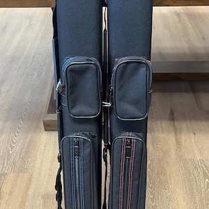 May include: Two black cue cases with contrasting stitching and zippered pockets. One case has blue stitching, the other red. Each case has a shoulder strap and a zippered compartment on the front.