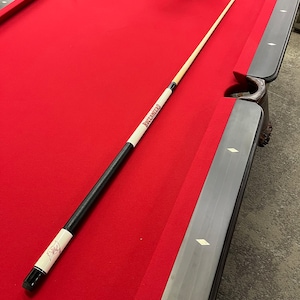 May include: A pool cue with a black leather grip and a white shaft rests on a red felt pool table. The cue features the word "BUCCANEERS" in red lettering on a white band. The table has dark wood trim.