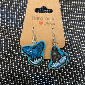 May include: A pair of handmade earrings featuring embroidered whale tails. One earring is blue with white accents, and the other is black with white accents. The earrings are on a brown card with the text "Handmade with love".