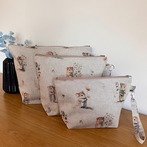 May include: Three beige linen zippered pouches with a floral and mouse print. The pouches are nested inside each other, with the largest pouch at the bottom and the smallest pouch at the top. The smallest pouch has a black zipper pull with a silver ring.