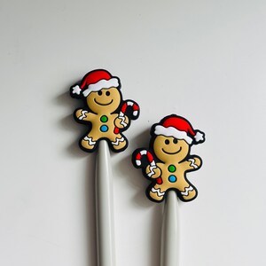 May include: Two gingerbread man-shaped Christmas ornaments with red and white Santa hats and candy cane accents. The ornaments are attached to gray pens.
