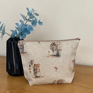 May include: A beige zippered pouch with a linen texture featuring a repeating pattern of brown mice holding flowers. The mice are surrounded by small floral details.