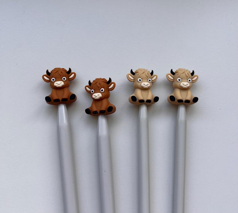 May include: Four knitting needles with animal-shaped tops. Two needles have brown cow tops and two needles have tan cow tops. The needles are silver.