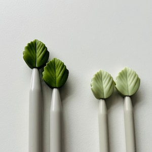 May include: Set of five knitting needles with leaf-shaped tops. Two needles have green leaves and three needles have light green leaves.