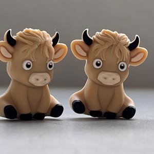 May include: Two brown and black cartoon cow figurines with white eyes and a light brown mane. The cows are sitting with their legs tucked under them.