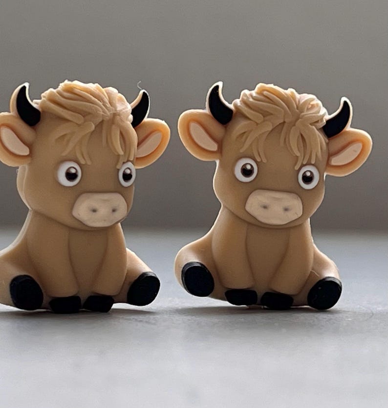 May include: Two brown and black cartoon cow figurines with white eyes and a light brown mane. The cows are sitting with their legs tucked under them.