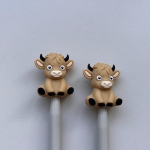 May include: Two knitting needles with brown cow-shaped toppers. The cows have black eyes, noses, and hooves.