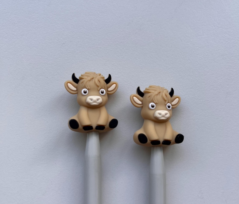 May include: Two knitting needles with brown cow-shaped toppers. The cows have black eyes, noses, and hooves.