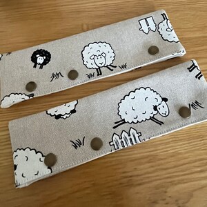 May include: Two beige fabric wristbands with a repeating pattern of black and white sheep. Each band has three bronze-colored snap closures. The sheep are depicted in various poses, some running and others grazing.
