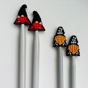 May include: Two pairs of knitting needles with gnome-themed toppers. One pair has red gnomes with black hats and the other pair has black gnomes with orange beards and skull hats.