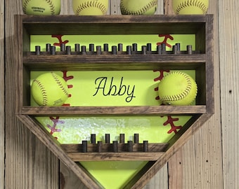 May include: A home plate-shaped wooden display shelf with a dark brown finish and a bright yellow back panel featuring red softball stitching graphics. The shelf has two horizontal tiers with small pegs for holding softballs, and the name Abby is printed in black script on the center back panel.