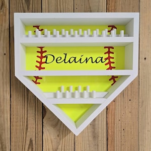 May include: A white home plate-shaped wall display shelf with two horizontal tiers featuring small white pegs for hanging items. The back panel is bright yellow with red softball stitching graphics and the black text Delaina printed centrally.
