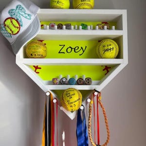 May include: A white baseball-shaped shelf with a neon yellow interior, displaying softballs, rings, and ribbons. The name "Zoey" is written on the shelf. A softball cap with a bow is on the left. Softballs have text on them.