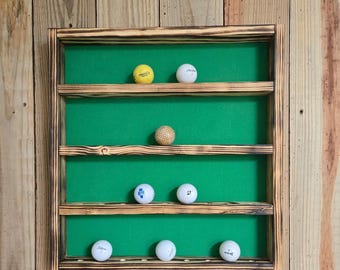 May include: A wooden golf ball display case with a green felt backing. The case has multiple shelves holding various golf balls. The wood has a dark, burnt finish. The golf balls are white, yellow, and patterned.