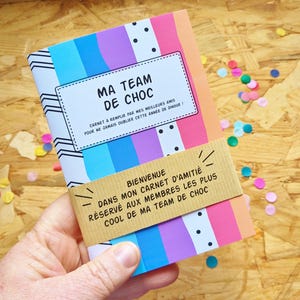 Peut inclure: Un carnet coloré avec une couverture rayée arc-en-ciel et le texte "MA TEAM DE CHOC" et "CARNET À REMPLIR PAR MES MEILLEURS AMIS POUR NE JAMAIS OUBLIER CETTE ANNÉE DE DINGUE!" sur la face avant. Le carnet est entouré d'une bande de papier brun avec le texte "BIENVENUE DANS MON CARNET D'AMITIÉ RÉSERVÉ AUX MEMBRES LES PLUS COOL DE MA TEAM DE CHOC".