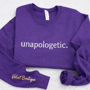 Puede incluir: Una sudadera morada con la palabra "unapologetic" en escritura blanca en el pecho. El puño de la manga presenta el texto bordado "Velvet Boutique" en dorado. La sudadera está hecha de un material suave.