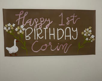May include: A brown paper banner with the words "Happy 1st Birthday Corin" written in pink and white paint. There are white flowers with green stems on either side of the text and a white goose with an orange beak and legs in the center.