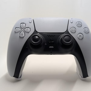 Könnte beinhalten: Ein kabelloser Videospiel-Controller in zweifarbigem Weiß und Schwarz. Das Gerät verfügt über ein Steuerkreuz, zwei schwarze Analogsticks, vier Aktionstasten mit geometrischen Symbolen, ein zentrales Touchpad und ein kleines PlayStation-Logo.
