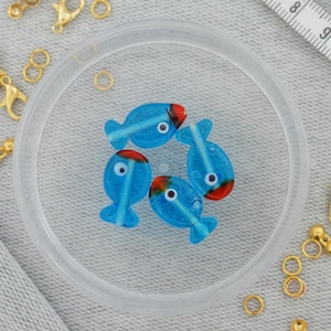 Könnte beinhalten: Fünf blaue Glasperlen in Fischform mit roten und weißen Akzenten. Die Perlen haben weiße Augen und sind in einem durchsichtigen Behälter kreisförmig angeordnet.