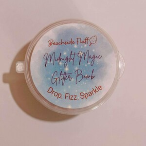 May include: A round, clear container with a blue and white label. The label reads "Beachside Fluff" and "Midnight Magic Glitter Bomb" with the words "Drop, Fizz, Sparkle" below. The container has a small tab on the side.