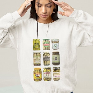 Peut inclure: Sweat-shirt blanc avec un imprim&eacute; graphique de neuf bocaux de cornichons diff&eacute;rents. Les bocaux sont dispos&eacute;s en trois rang&eacute;es de trois. Chaque bocal porte une marque diff&eacute;rente et un type de cornichon diff&eacute;rent. Le sweat-shirt est de couleur gris clair et a un col rond.