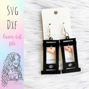 May include: A pair of black guillotine-shaped earrings with a rose gold blade. The earrings are hanging from silver hooks.
