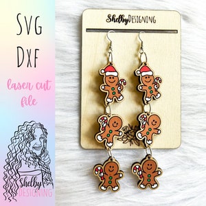 May include: A pair of gingerbread man dangle earrings with red, green, and white details. The earrings are made of wood and have a silver hook.
