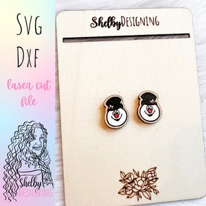 May include: A pair of wooden snowman earrings with black hats and red noses. The earrings are displayed on a wooden board with the text "ShelbyDESIGNING" on it.