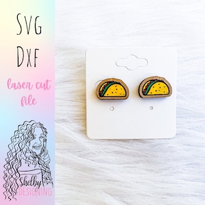 May include: A pair of wooden taco earrings. The earrings are painted yellow with green, red, and brown details. The earrings are on a white card.