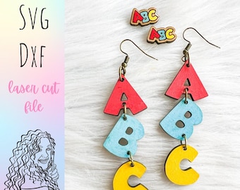 Op de afbeelding: Een paar hangende oorbellen met houten letters A, B en C. De letters zijn rood, blauw en geel geverfd. De oorbellen hangen aan gouden haken.
