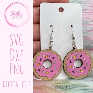May include: A pair of pink donut-shaped earrings with white and purple sprinkles. The earrings are made of wood and have silver hooks.