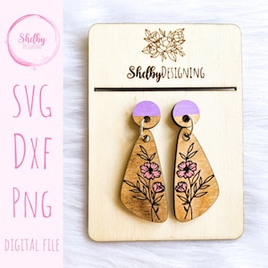 Puede incluir: Un par de pendientes de madera con un diseño floral rosa y una parte superior morada. Los pendientes se muestran en un soporte de madera con el texto "Shelby DESIGNING" en él.