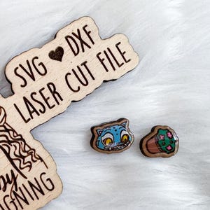 May include: Wooden laser cut file with a woman's portrait and the text "Shelby DESIGNING". Two small, colorful wooden earrings are next to the file. One earring is a blue cat, and the other is a green cactus.