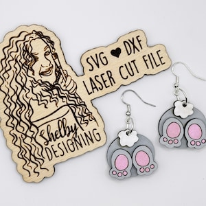 May include: A pair of laser cut wooden earrings shaped like bunny bottoms. The earrings are gray with pink accents and have a white flower charm hanging from the top. The earrings are packaged with a wooden sign that says "SVG ♥ DXF LASER CUT FILE" and "Shelby Designing".