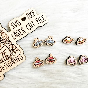 May include: A set of six laser cut wooden earrings featuring different colored UFO designs. The earrings are displayed on a wooden sign that reads "SVG ♥ DXF LASER CUT FILE Shelby Designing".