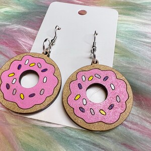 May include: A pair of pink donut earrings with white and purple sprinkles. The earrings are made of wood and have silver hooks.