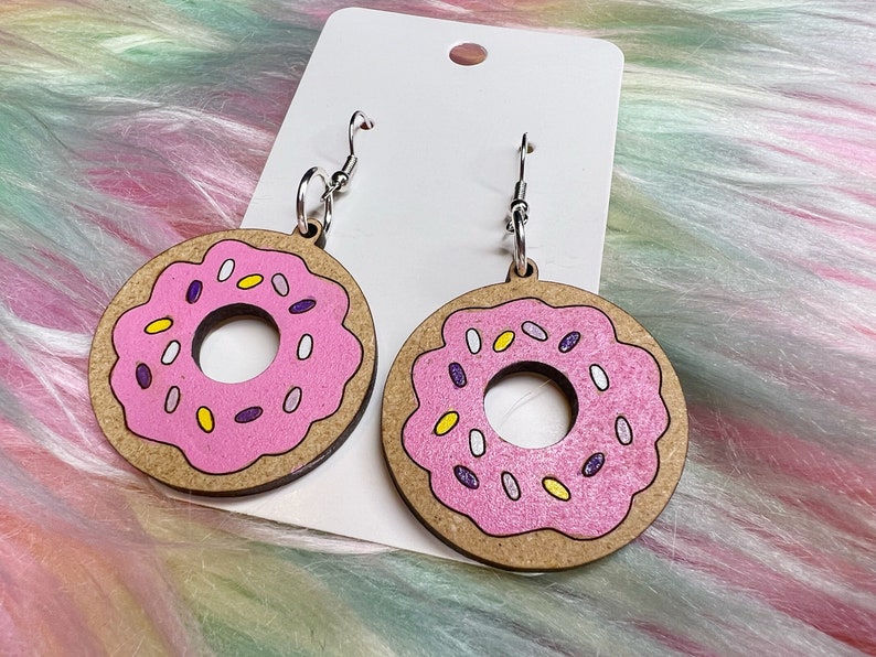 May include: A pair of pink donut earrings with white and purple sprinkles. The earrings are made of wood and have silver hooks.